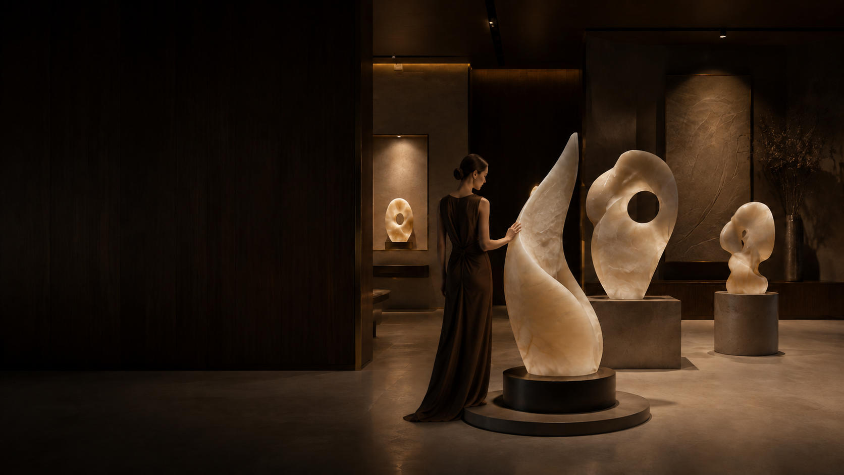Backlit alabaster sculptures in a dimly lit private studio gallery, with a woman studying one of the forms